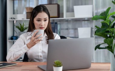 Modern ofis masasındaki Asyalı hevesli genç iş kadını bir fincan kahveyle çalışmak için dizüstü bilgisayar kullanıyor. Çalışkan ve çekici ofis hanımı ofisinde bilgisayar defteri üzerinde çalışıyor..
