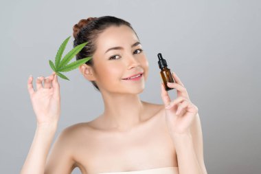 Glamorous beauty portrait of beautiful young woman with flawless clean skin cosmetic makeup holding cannabis green leaf, CBD extracted oil bottle for natural skincare treatment in isolated background.