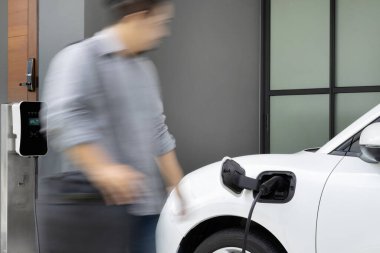 Focus electric car charging at home charging station with blurred progressive man walking in the background. Electric car using renewable clean for eco-friendly concept.