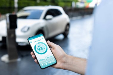 Closeup progressive businessman look at EV cars battery status application on smart phone screen at public parking charging station with power cable plug and renewable energy-powered electric vehicle