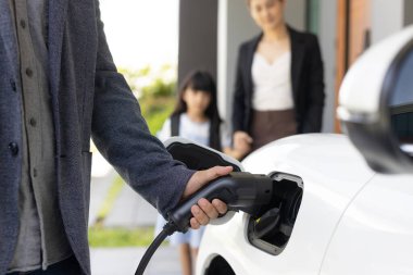 Elektrikli arabaya odaklan, elektrikli araca şarj cihazı yerleştir. Arka planda bulanık aile ile alternatif yeşil enerji teknolojisi kavramını geliştir..