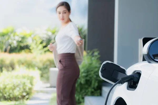 Focus EV charger plugged into EV car at home charging station with blurred background of progressive woman walking in background. Elective vehicle powered by clean energy for eco-friendly concept.