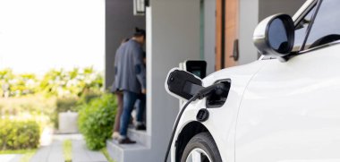 Focus closeup electric vehicle recharging battery from home electric charging station with blurred family in background. Renewable clean energy car for progressive eco awareness lifestyle concept.