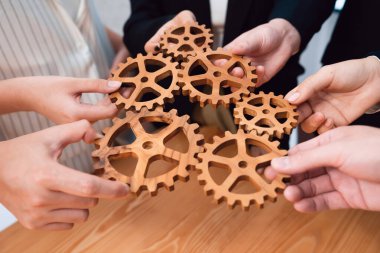 Closeup hand holding wooden gear by businesspeople wearing suit for harmony synergy in office workplace concept. Group of people hand making chain of gears into collective form for unity symbol.