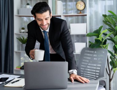 Modern ofis masasındaki modern profesyonel iş adamı elinde kahveyle çalışmak için dizüstü bilgisayar kullanıyor. Ofisinde bilgisayar defteri üzerinde çalışan çalışkan bir ofis çalışanı. coşkulu