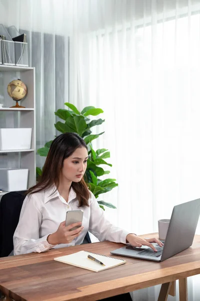 現代的で若い熱心なビジネス女性のマルチタスクは ビジネス上の問題のために彼女のスマートフォンをチェックし オフィスで彼女の机の上でノートパソコンで作業することによって — ストック写真