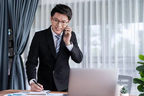 Businessman working on laptop for on office desk workspace with his smartphone. Smart executive researching financial business data or planning strategic business, talking on phone. Jubilant