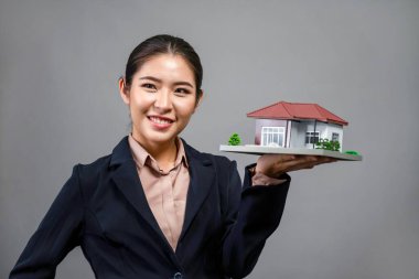 Resmi takım elbise giyen genç Asyalı kadın izole edilmiş arka planda ev modelini tutuyor ve sergiliyor. Konut ilanı, ev sigortası promosyonları ya da kişiselleştirilebilir fotokopi alanı için ideal. Coşkulu.