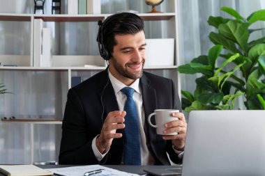 Çağrı merkezi operatörünün müdürü bilgisayarla uğraşırken masasında kahvesiyle oturuyor. Kulaklık takan ve siyah takım giyen bir ofis çalışanı müşteri desteği ya da tele pazarlama üzerinde çalışıyor. coşkulu