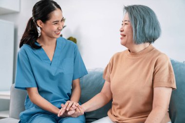 A caregiver rest her hands on the shoulders of a contented senior patient while she sitting on the sofa at home.