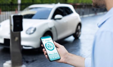 Closeup progressive businessman look at EV cars battery status application on smart phone screen at public parking charging station with power cable plug and renewable energy-powered electric vehicle