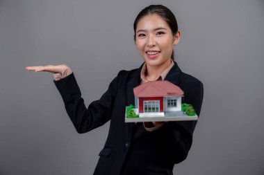 Resmi takım elbise giyen genç Asyalı kadın izole edilmiş arka planda ev modelini tutuyor ve sergiliyor. Konut ilanı, ev sigortası promosyonları ya da kişiselleştirilebilir fotokopi alanı için ideal. Coşkulu.