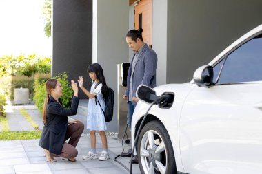 Progressive young parents and daughter with electric vehicle and home charging station. Green and clean energy from electric vehicles for healthy environment. Eco power from renewable source at home.