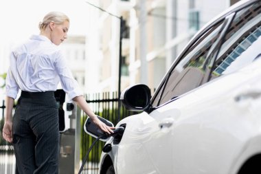 Progressive businesswoman insert charger plug from charging station to her electric vehicle with apartment condo building in background. Eco friendly rechargeable car powered by sustainable energy.