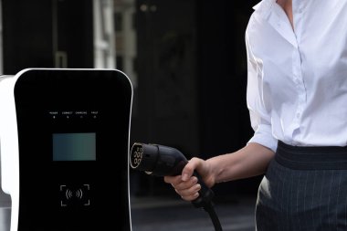 Focus EV charger plug and electric car at public charging station with blur progressive businesswoman holding charger and apartment condo building in background. Eco friendly electric vehicle concept.