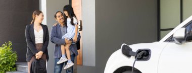 Progressive young parents and daughter with electric vehicle and home charging station. Green and clean energy from electric vehicles for healthy environment. Eco power from renewable source at home.
