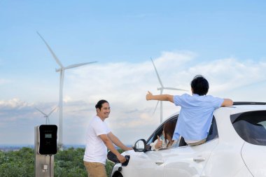 İlerici mutlu aile kavramı elektrikli araçla rüzgar çiftliğinde eğleniyor. Rüzgar türbini jeneratöründen gelen temiz yenilenebilir enerji ile şarj edilen elektrikli araç..