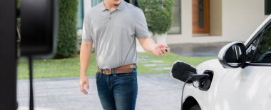 Progressive asian man install cable plug to his electric car with home charging station in the backyard. Concept use of electric vehicles in a progressive lifestyle contributes to clean environment.