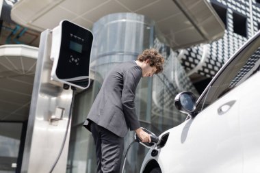 Below view closeup image of progressive black suit businessman recharge battery of his electric vehicle from public charging station. Renewable and alternative energy powered car concept.