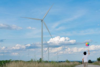 Progressive young asian boy playing with wind pinwheel toy in the wind turbine farm, green field over the hill. Green energy from renewable electric wind generator. Windmill in the countryside concept