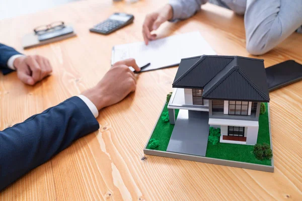 House model is displayed on wooden meeting table with in the blurred background of real estate agent and client discuss terms and conditions of house loan or rental lease contract. Entity