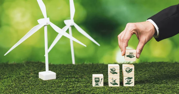 Businessman putting Net Zero symbol cube on stack top for corporate use eco-friendly energy and sustainability. Wind turbine generate renewable and clean energy with net zero CO2 emission. Alter