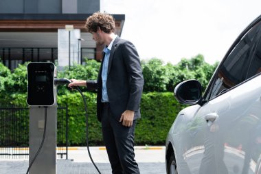 Modern şehir yerleşim bölgesindeki kamu şarj istasyonunda elektrikli araba şarj eden yenilikçi bir işadamı. Çevre dostu şarj edilebilir araba alternatif temiz enerjiyle çalışıyor..