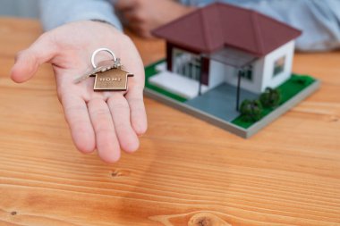 Emlakçı, ev anahtarını hesap makinesi ve yasal belgelerle çevrili ahşap masanın üzerinde, ikna edici ev satışları ve ipotek teklifinin sembolik bir jesti olarak sunuyor. Varlık