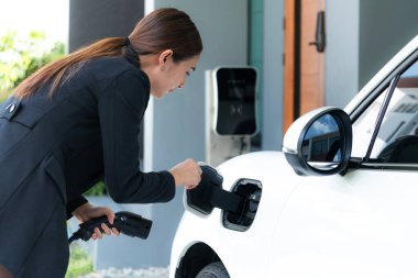 Progressive woman install cable plug to her electric car with home charging station. Concept of the use of electric vehicles in a progressive lifestyle contributes to clean environment.