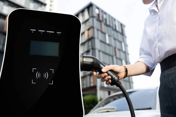 Focus EV charger plug and electric car at public charging station with blur progressive businesswoman holding charger and apartment condo building in background. Eco friendly electric vehicle concept.
