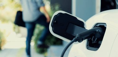 Focus electric car charging at home charging station with blurred progressive man walking in the background. Electric car using renewable clean for eco-friendly concept.