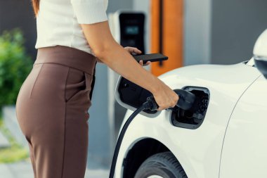 İlerici kadın elektrikli arabasının fişini şarj istasyonuna takıyor. İlerici bir yaşam tarzında elektrikli araç kullanımı kavramı temiz çevreye katkıda bulunur.