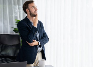 Siyah takım elbiseli yakışıklı iş adamı, modern ofis portresine güvenerek duruyor. Düşünceli bir ifadeyle iş hakkında derin düşüncelere dalmış bir sonraki hamlesini stratejik olarak düşünüyor. Varlık