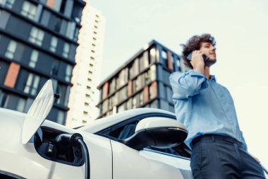 Progressive businessman talking on the phone, leaning on electric car recharging with public EV charging station, apartment condo residential building on the background as green city lifestyle.