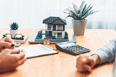 Ev modeli ahşap toplantı masasında, emlakçı ve müşterinin bulanık geçmişinde ev kredisi veya kira kontratının şartlarını tartışırken görüntülenir. Varlık
