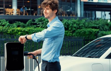 Businessman with smartwatch at modern charging station for electric vehicle with background of residential buildings as concept for progressive lifestyle of using eco-friendly as alternative energy.