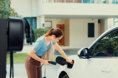 Progressive woman install cable plug to her electric car with home charging station. Concept of the use of electric vehicles in a progressive lifestyle contributes to clean environment.