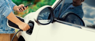 Progressive natural scenic where hand insert charging plug to electric vehicle from charging station with natural background. Future energy concept of EV car powered by sustainable electric energy.