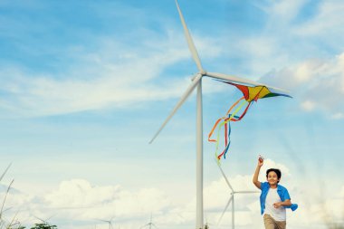 İlerici mutlu kaygısız çocuk konsepti. Yol boyunca koşan ve uçurtma uçuran genç bir çocuk kırsal alanda doğal manzaralı, açık gökyüzü ve güneşli bir gün dağ ve rüzgar türbini arka planında