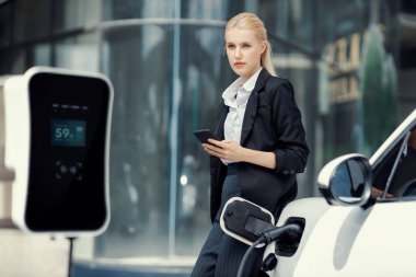 Businesswoman wearing black suit using smartphone, leaning on electric car recharge battery at charging station in city residential building with condos and apartment. Progressive lifestyle concept.