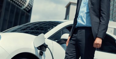 Closeup progressive businessman with electric car recharging at public charging station with background of city residential building. Eco friendly car powered by alternative clean energy.