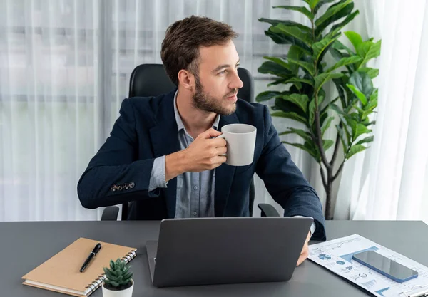 stock image Businessman working on his laptop and drinking coffee at his desk in workspace. Smart executive taking coffee break after researching financial data and marketing plan on his corporate laptop. Entity