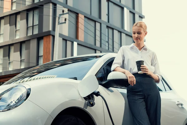 Businesswoman Drinking Coffee Leaning Electric Vehicle Recharging Public Charging Station — Stok fotoğraf