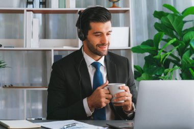 Çağrı merkezi operatörünün müdürü bilgisayarla uğraşırken masasında kahvesiyle oturuyor. Kulaklık takan ve siyah takım giyen bir ofis çalışanı müşteri desteği ya da tele pazarlama üzerinde çalışıyor. coşkulu