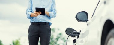 Closeup progressive suit-clad businessman with his electric vehicle recharge his car on public charging station in modern city with power cable plug and renewable energy-powered electric vehicle.