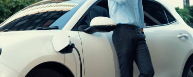 Closeup progressive suit-clad businessman with his electric vehicle recharge his car on public charging station in modern city with power cable plug and renewable energy-powered electric vehicle.