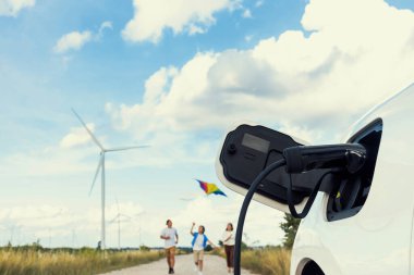 Concept of progressive happy family holding windmill toy and relax at wind farm with electric vehicle. Electric vehicle driven by clean renewable energy from wind turbine generator to charger station.