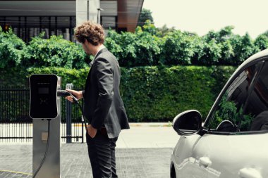 Modern şehir yerleşim bölgesindeki kamu şarj istasyonunda elektrikli araba şarj eden yenilikçi bir işadamı. Çevre dostu şarj edilebilir araba alternatif temiz enerjiyle çalışıyor..