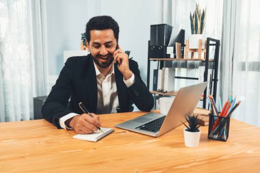 Çalışkan işadamı, ofis masasında dizüstü bilgisayarla çalışırken müşterileriyle telefonda konuşmakla meşgul. Cep telefonlu, modern, çalışkan ofis çalışanı yaşam tarzı kavramı olarak. Coşkulu