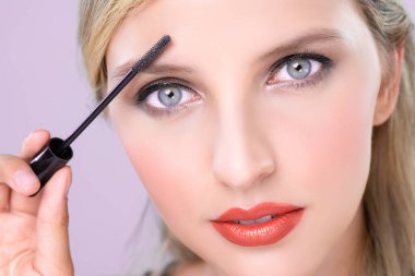 Closeup woman with blond hair putting alluring black mascara with brush in hand on long thick eyelash. Perfect fashionable cosmetic clean facial skin with beautiful eye young woman in high resolution.